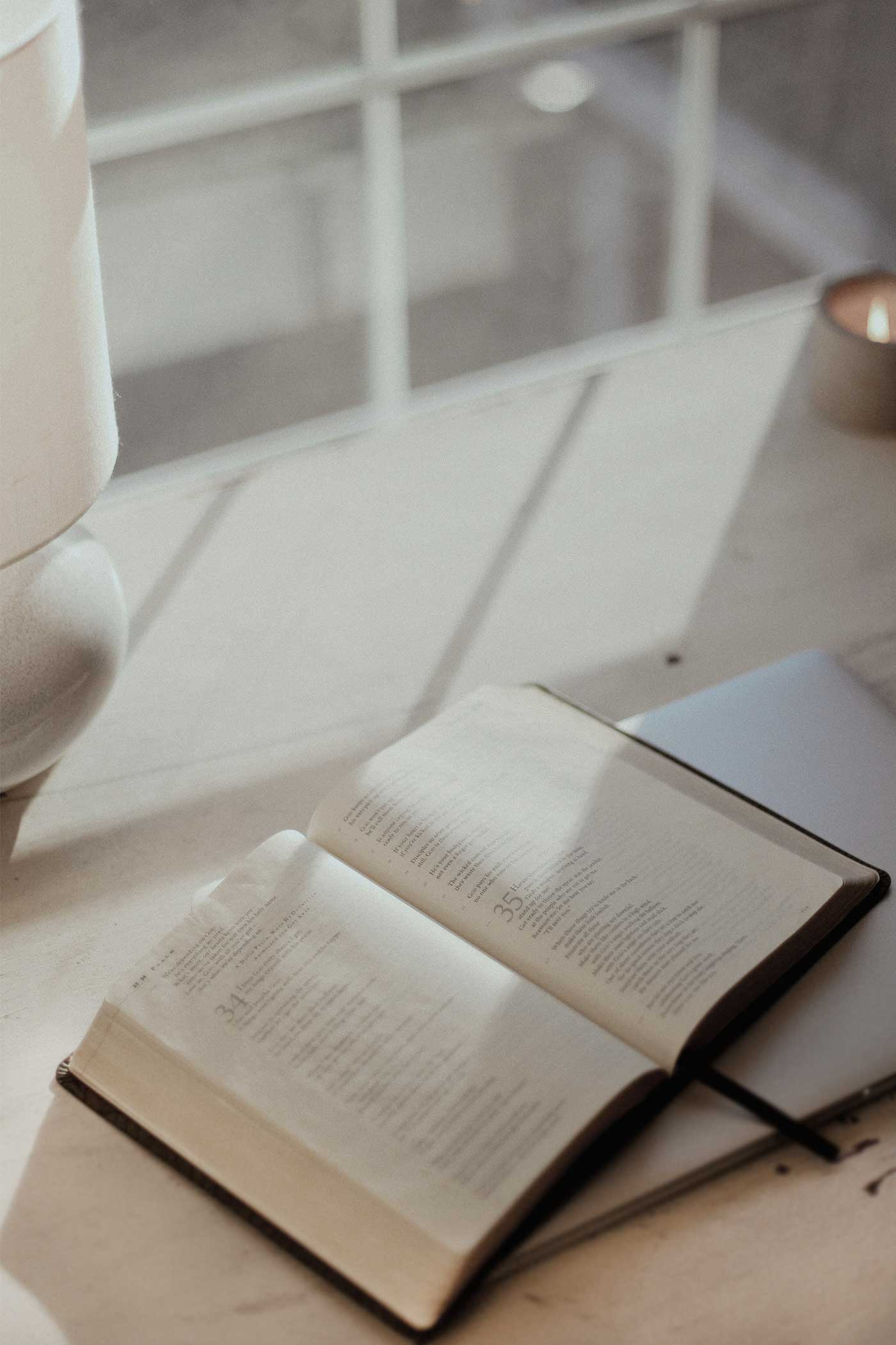 Open Bible resting on a notebook beside a window, with sunlight across the pages and a candle nearby.