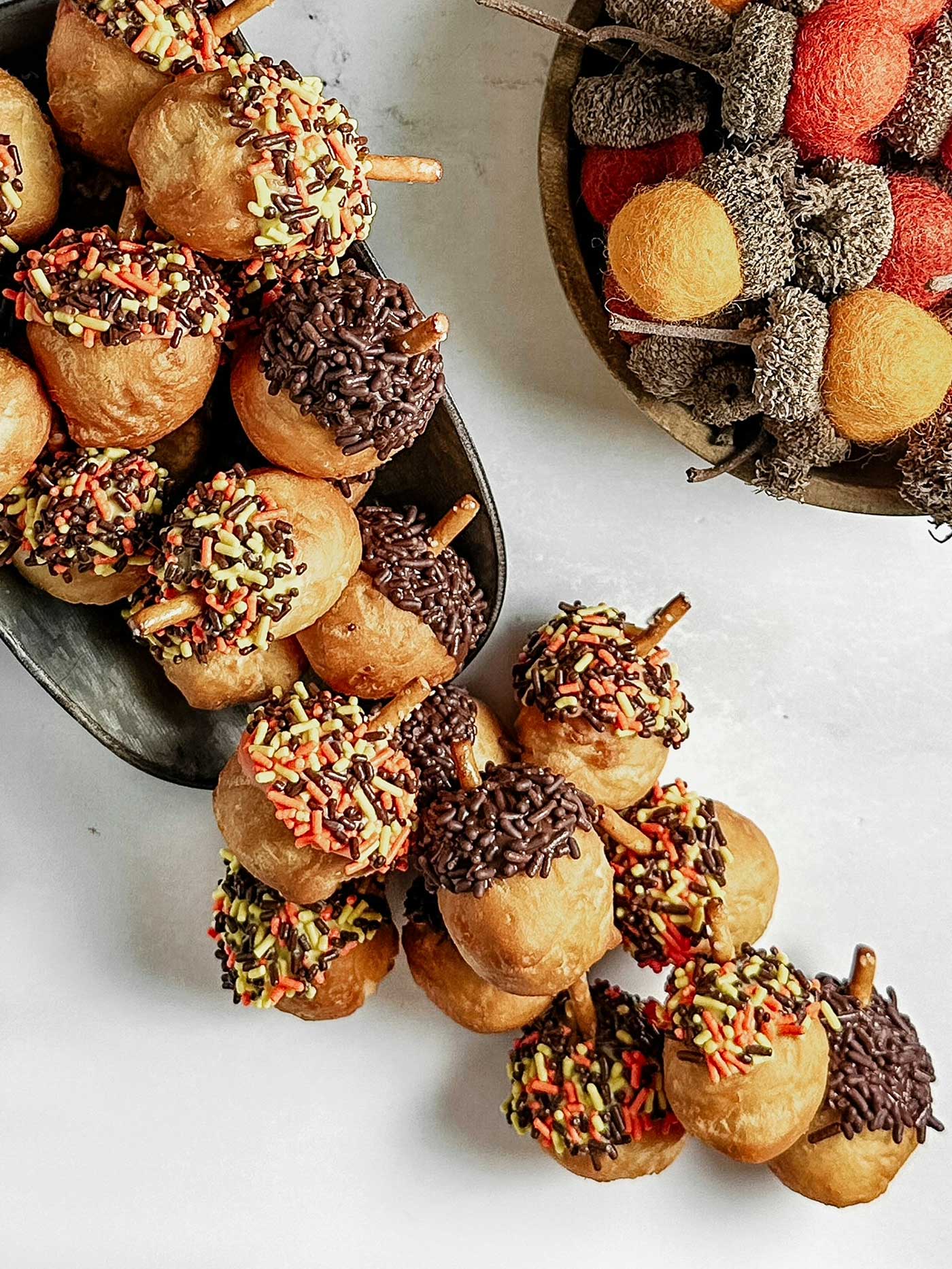 Small golden-brown acorn-shaped treats topped with chocolate and colorful fall sprinkles, with pretzel sticks for stems.